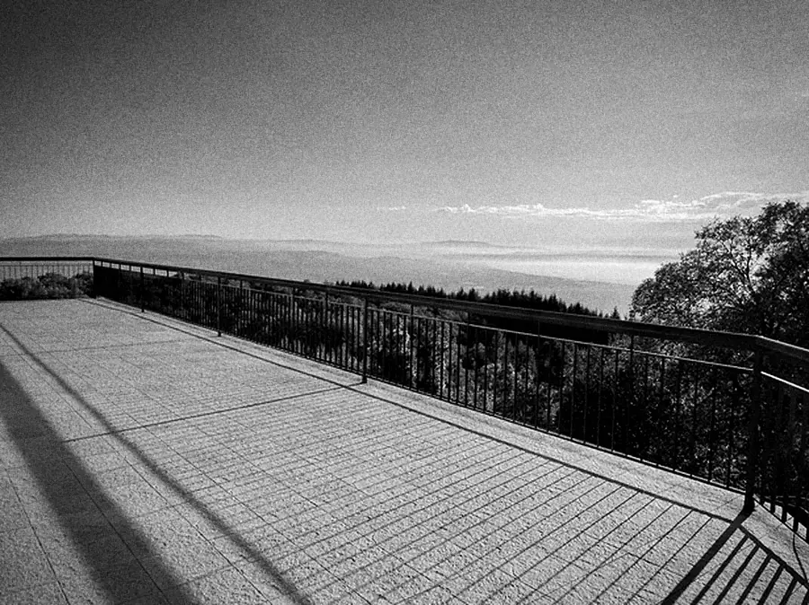 A wide terrace with a railing, looking over wooded hills towards the sea.